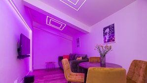 A modern living room with a purple-lit ceiling, a flat-screen TV mounted on the wall, a round dining table with yellow chairs, and a sofa with a floral vase. - ShortStayPoland Zelazna B146 (Warsaw)