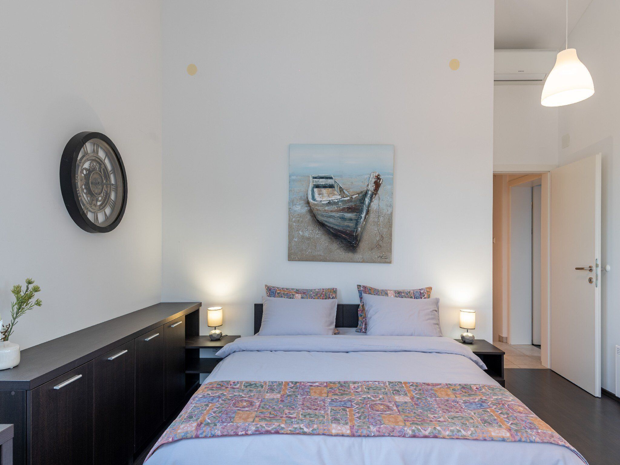 A bedroom with a bed, a nightstand, a wall clock, a painting of a boat, and a door.