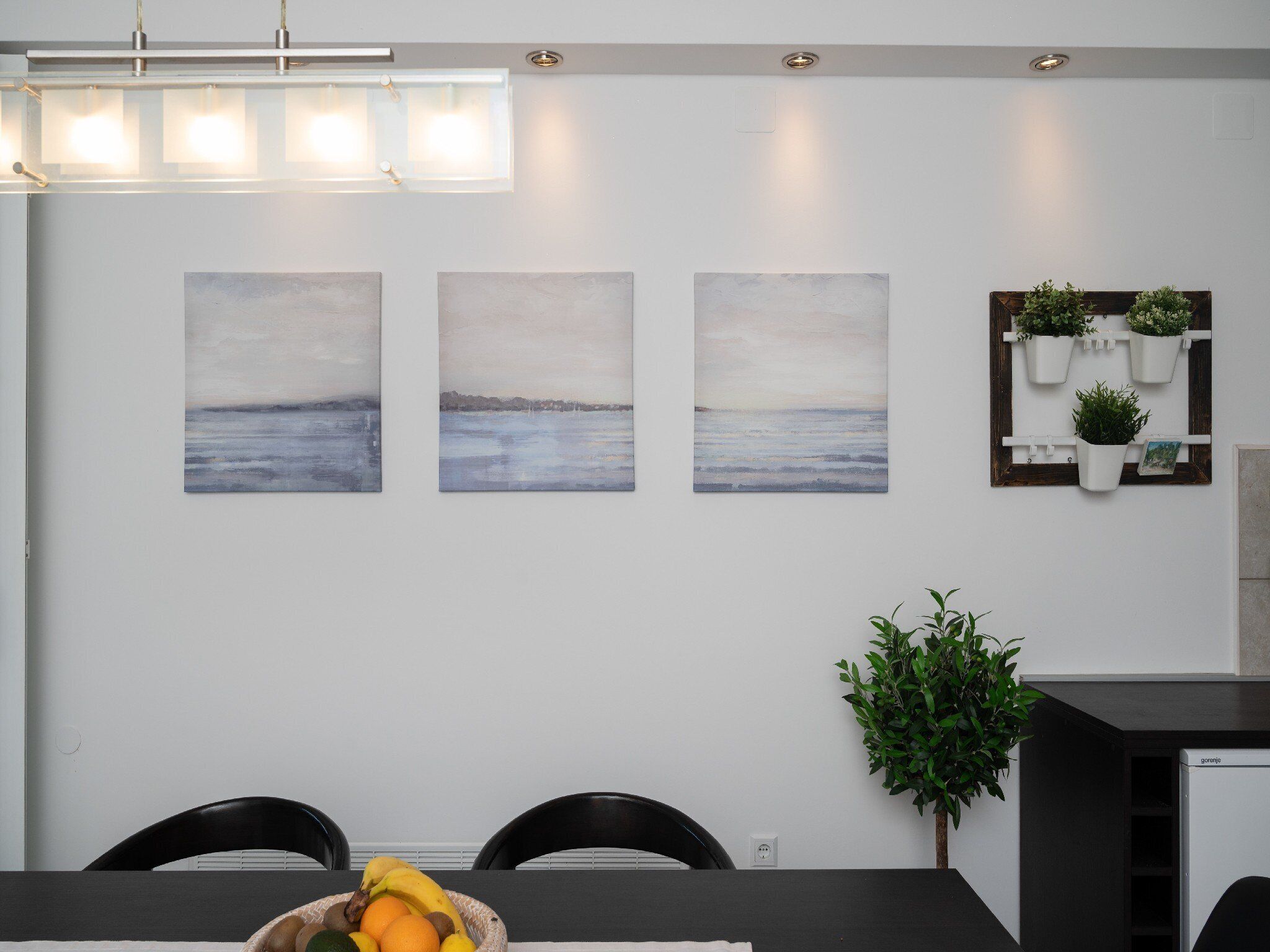 A modern dining area with a dark table, three wall-mounted paintings of a seascape, a small plant shelf, and a potted plant.