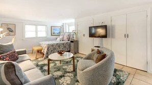 Una sala de estar con sofá, sillón, mesa de centro, televisión y una cama con colcha estampada.