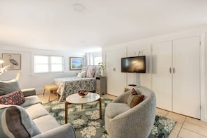 A modern living room with a sofa, armchair, and a small coffee table. There is a bed with a floral bedspread in the background, a television mounted on the wall, and a large wardrobe.