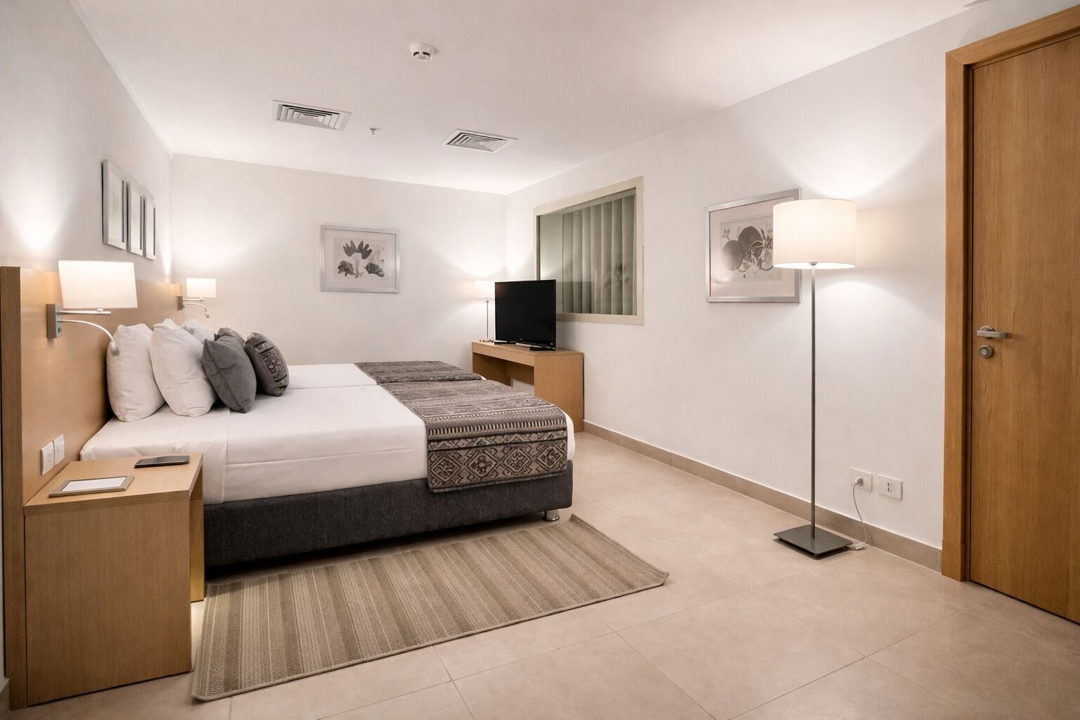 A modern hotel room with a bed, bedside tables, a flat-screen TV, a standing lamp, and a door.