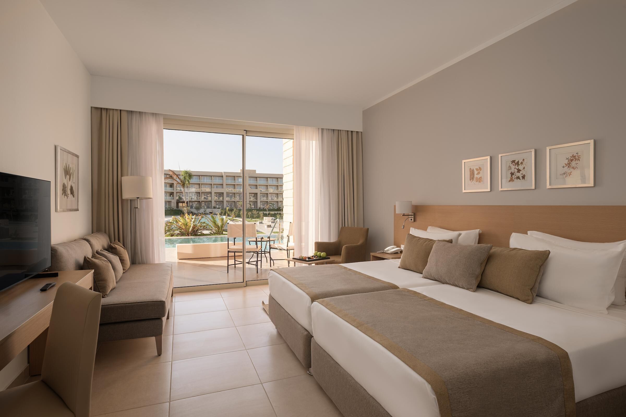 A hotel room with a bed, a desk, a TV, a chair, a lamp, and a view of a pool and building.