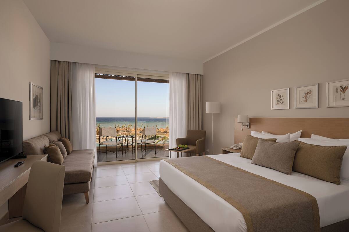 A hotel room with a large bed, a TV, a desk, and a balcony with a view of the beach.