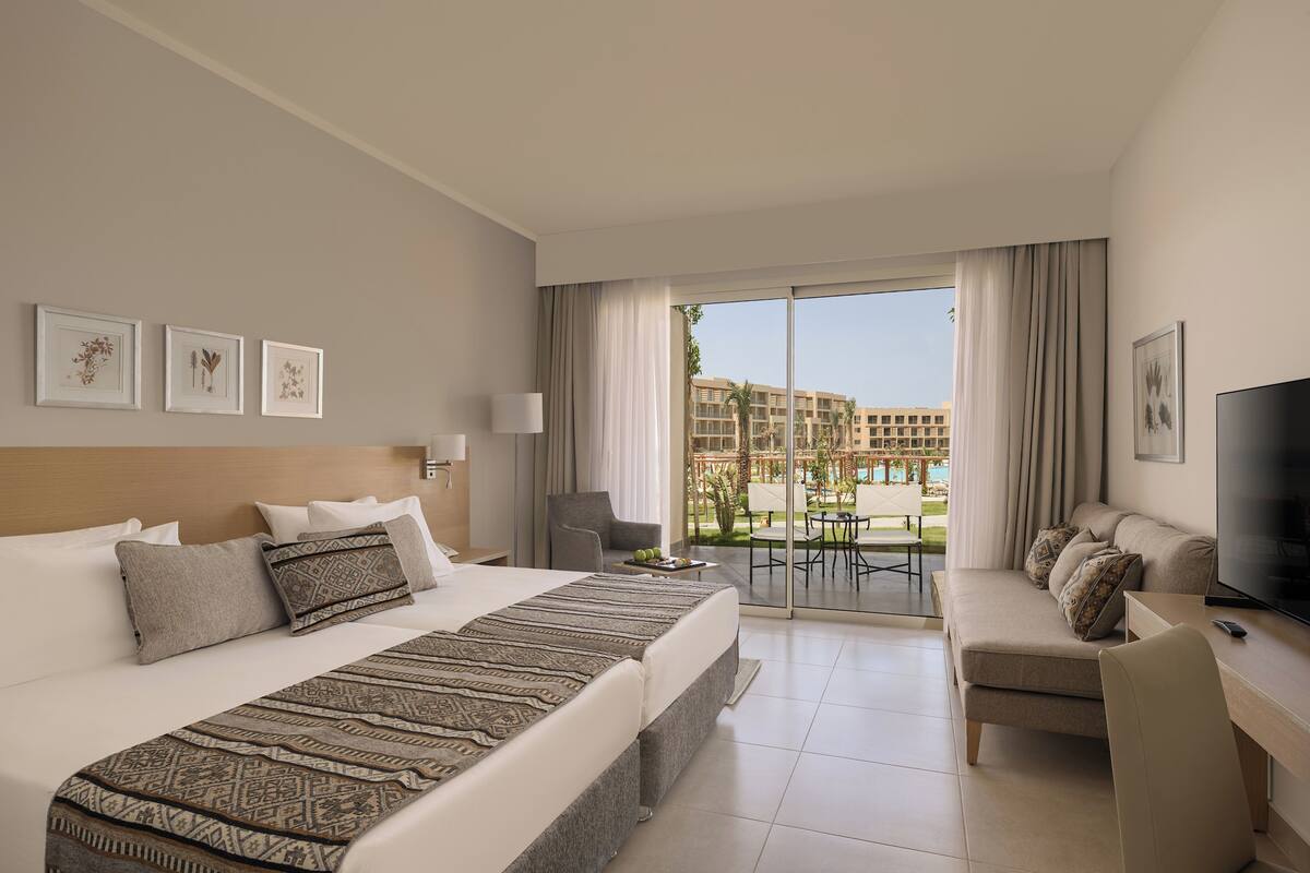 A hotel room with a large bed, a desk with a TV, a chair, and a balcony with a view of a building and greenery.