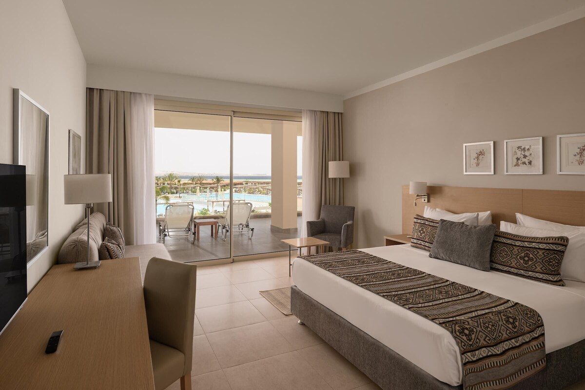 Una habitación de hotel con una cama, un escritorio, una silla, un televisor y vista a la playa.