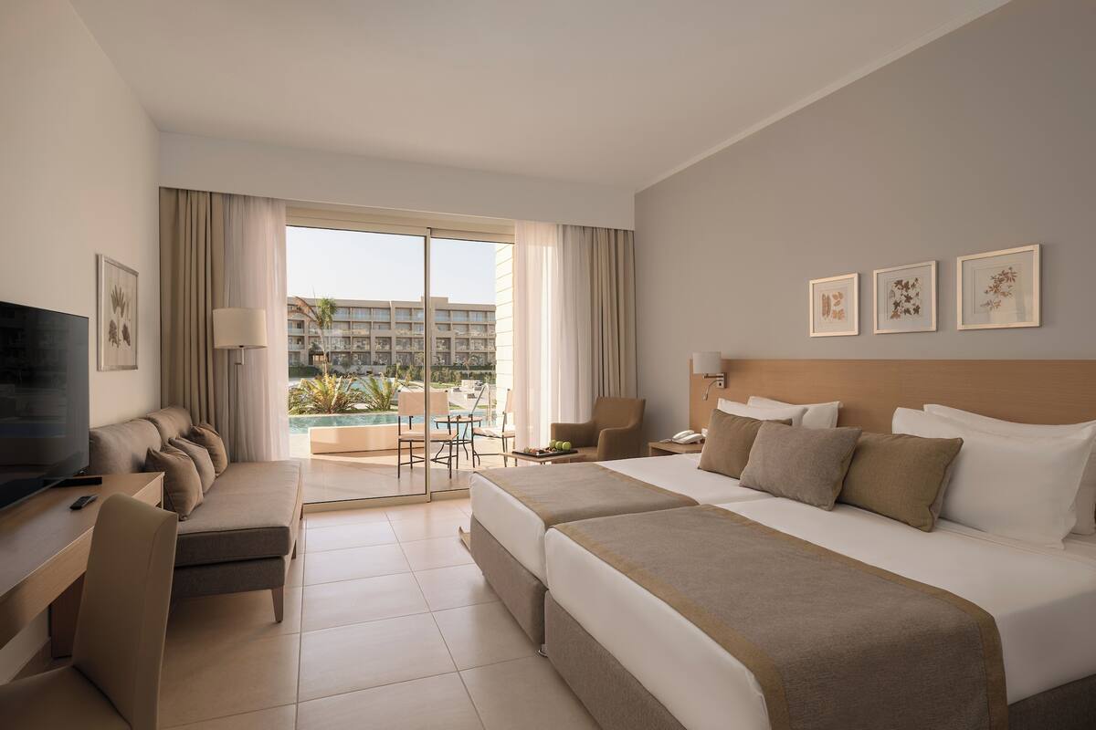A hotel room with a bed, a desk, a TV, a chair, a lamp, and a view of a pool and building.