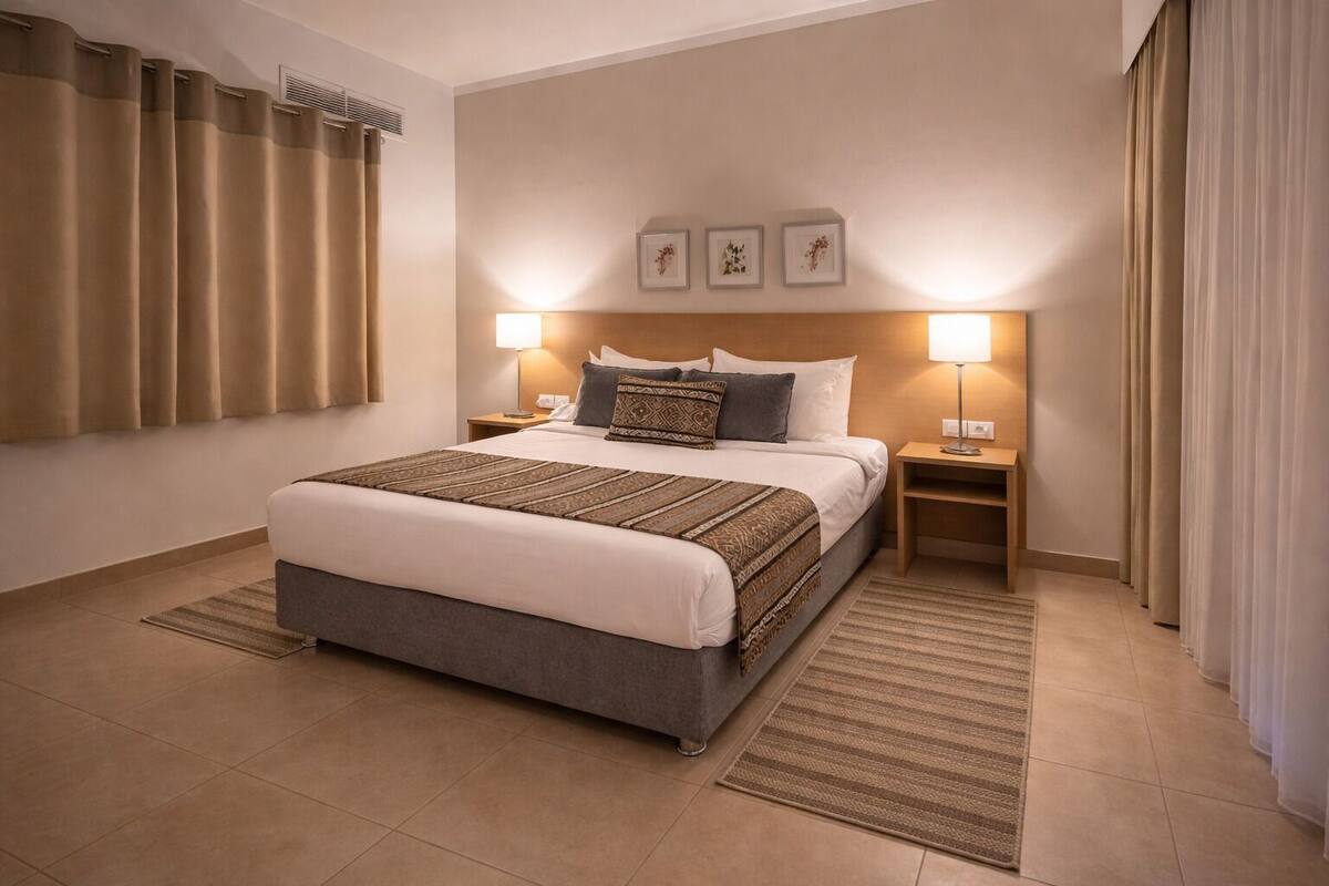 A hotel room with a bed, two bedside tables, a headboard, a wall-mounted air vent, and a window with curtains.