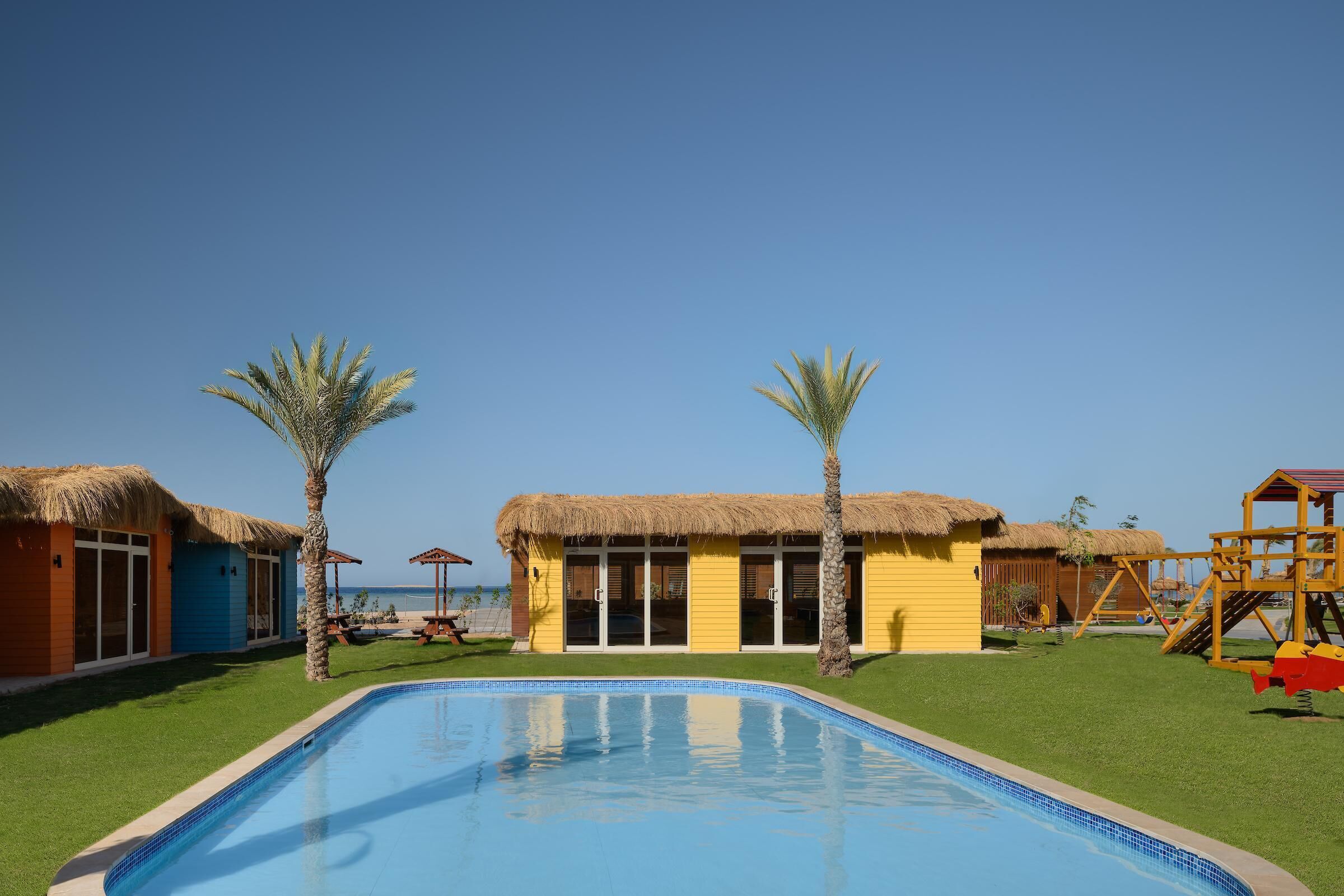 Un área de piscina con vista despejada al océano, un edificio con techo de paja y un parque infantil.