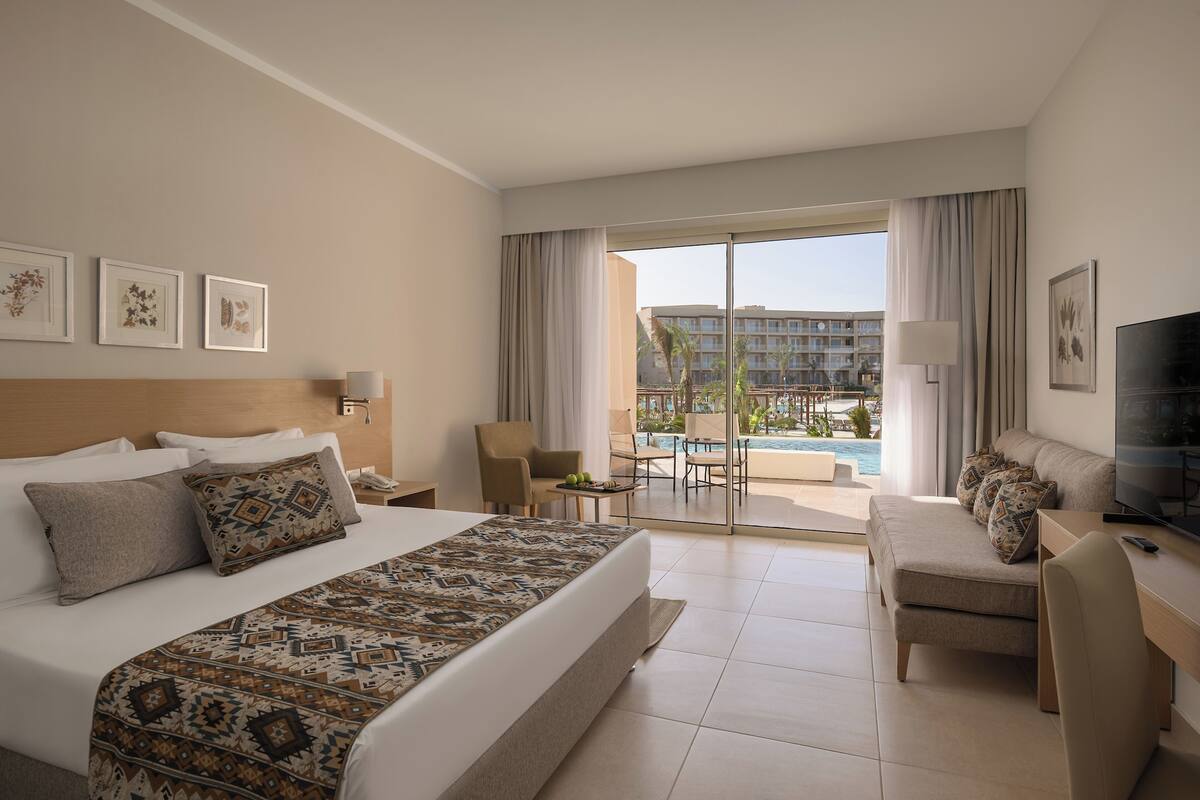 A hotel room with a large bed, a sofa, a TV, and a view of a swimming pool and building.