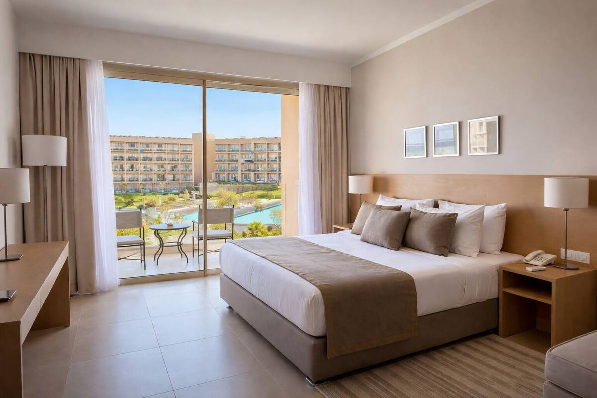 A hotel room with a bed, bedside tables, a desk, a chair, and a view of a pool and buildings.