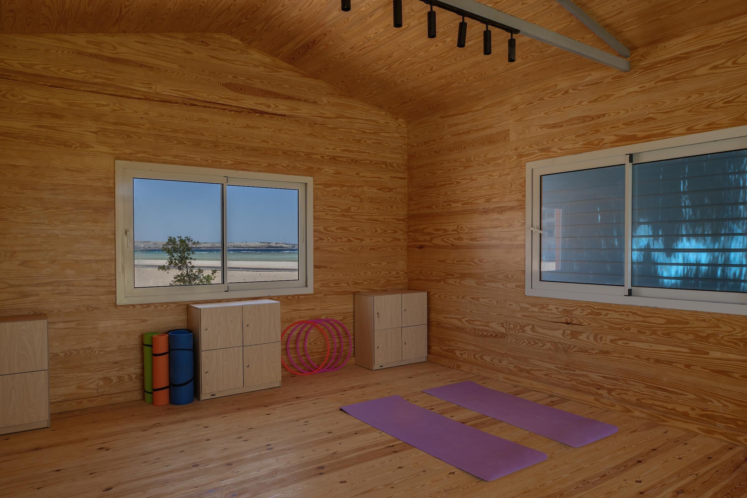 Una habitación con paredes de madera, dos ventanas, una lámpara de techo, un armario y colchonetas de yoga.