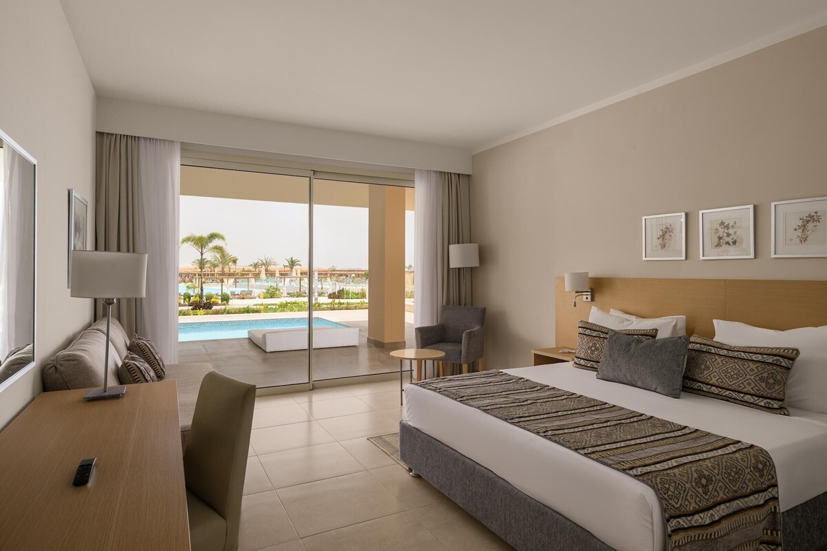 Habitación de hotel con cama, escritorio, silla, lámpara y vistas a una piscina y palmeras.