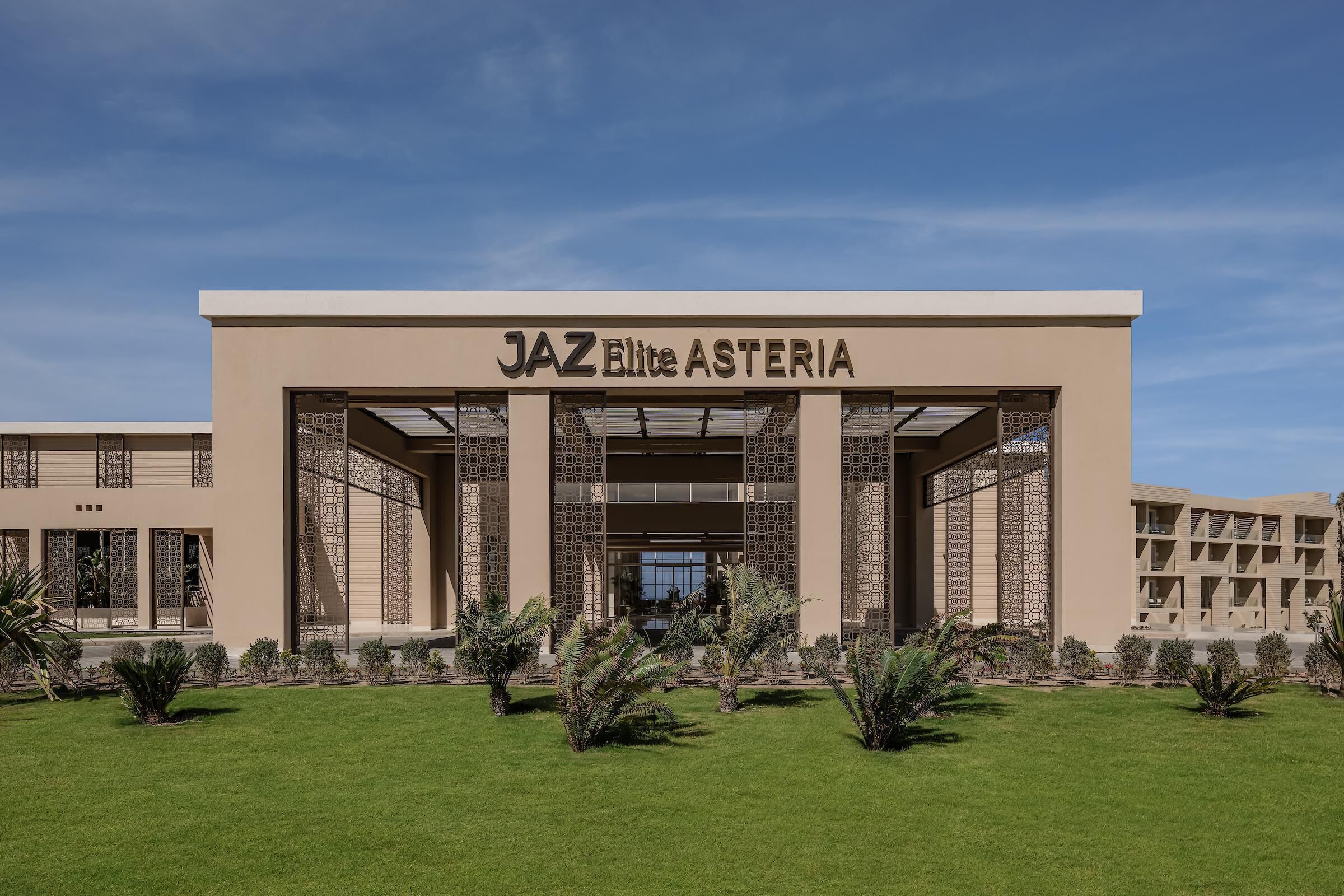 A building with a sign reading 'JAZ Elite Asteria' and a well-maintained lawn with small trees.