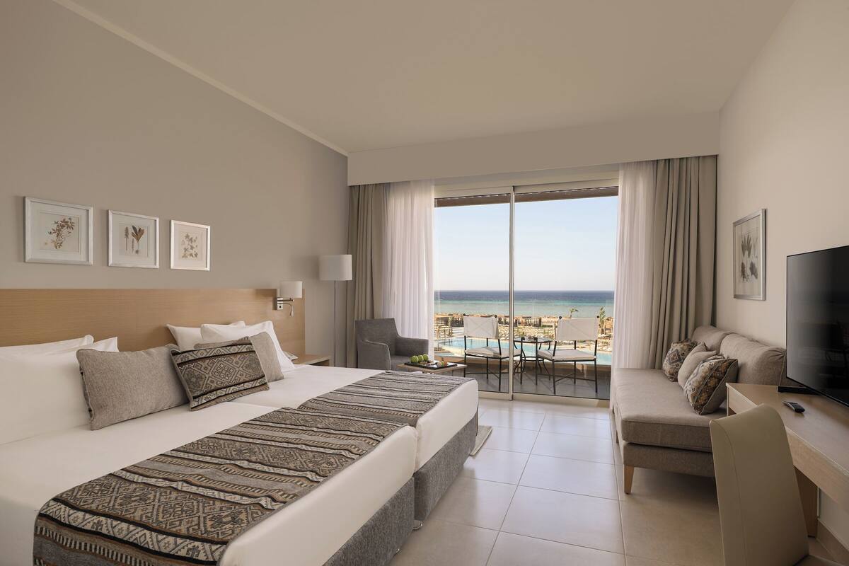A hotel room with a large bed, a desk with a TV, a balcony with a pool view, and framed artwork on the wall.