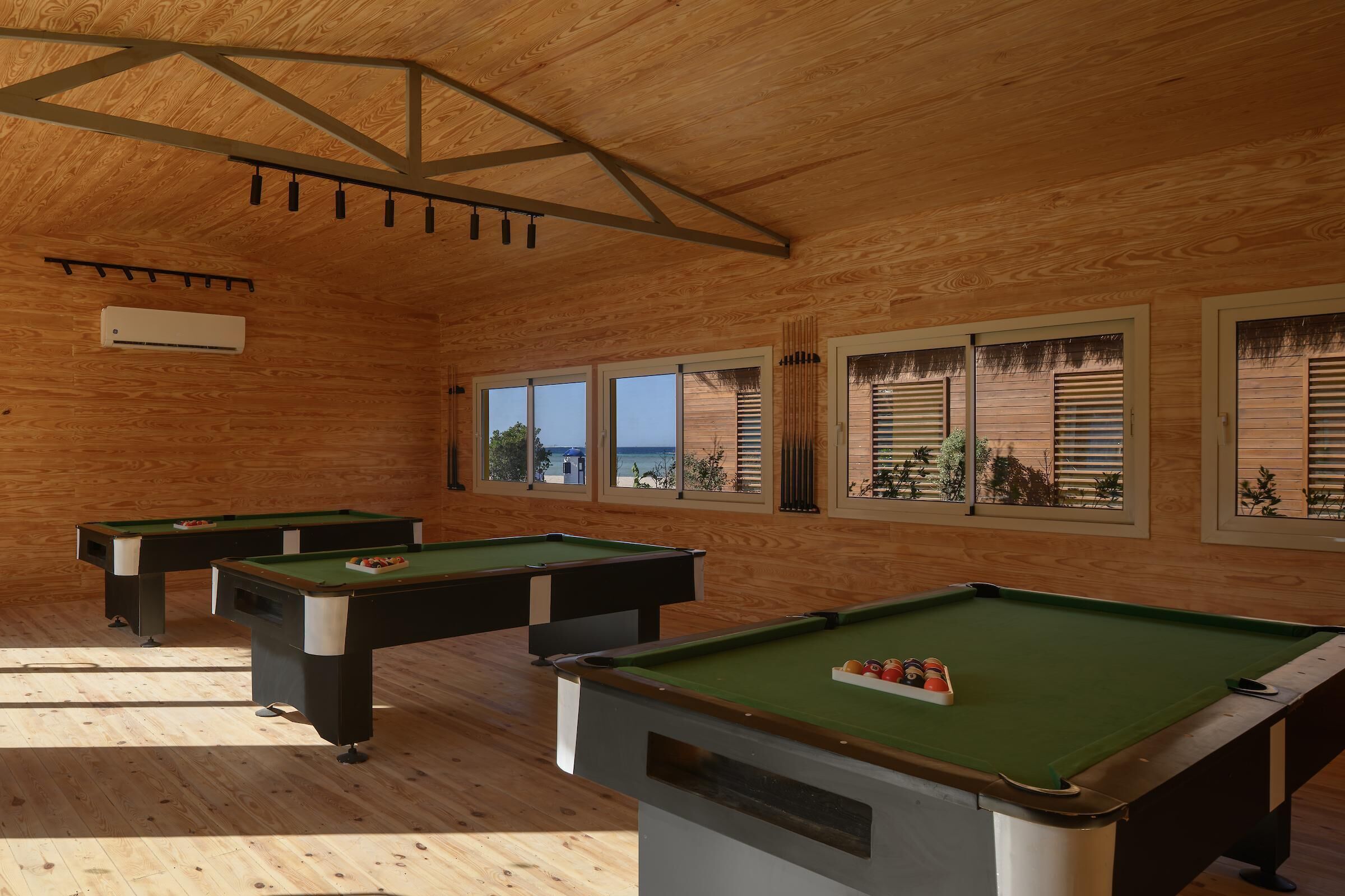 Une salle de billard aux murs et au plafond en bois, avec de nombreuses tables de billard et de grandes fenêtres offrant une vue dégagée.