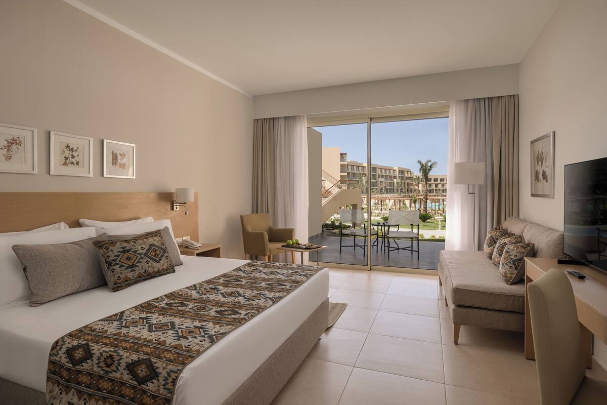 A hotel room with a large bed, a sofa, a TV, and a balcony with a view of buildings and a pool.