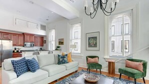 Una sala de estar moderna con sofá, reposapiés y dos sillones, una cocina con armarios de madera y ventanas amplias con persianas.