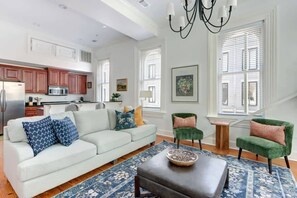 A spacious living area with a sofa, ottoman, and chairs, a kitchen with wooden cabinets, and large windows with blinds.
