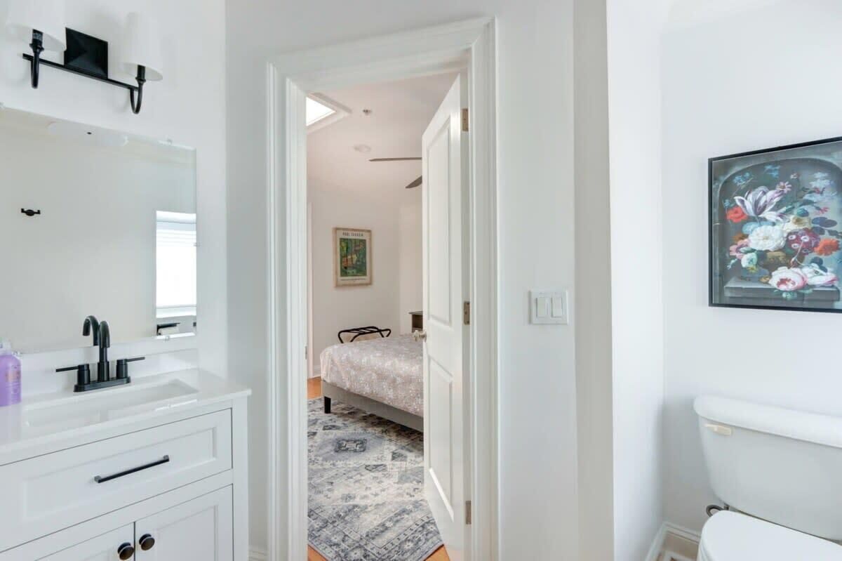 A bedroom with a bed, a white wall, a painting, and a bathroom with a sink and toilet.