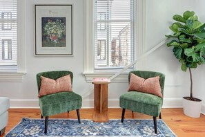 A room with two green armchairs, a blue patterned rug, a wooden side table, a framed artwork, and a potted plant.
