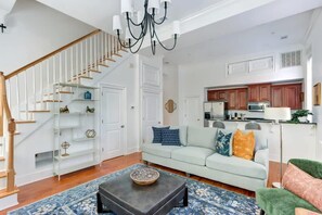 A living room with a staircase, a light-colored sofa, a coffee table, and a kitchen area with wooden cabinets and a microwave.
