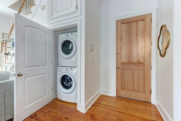 A laundry room with two stacked washing machines, a wooden door, and a white wall with a gold-framed mirror.