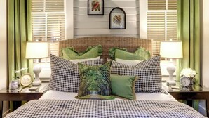 A bed with checkered bedding, green pillows, a wicker headboard, and two wall-mounted picture frames. - Avery's Cottage @ Trustees' Garden | Walk 2 River! (Savannah)