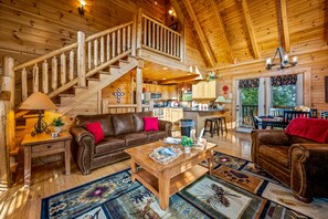 Amplia cabaña de madera con zona de estar, cocina y escalera que lleva a un nivel superior.