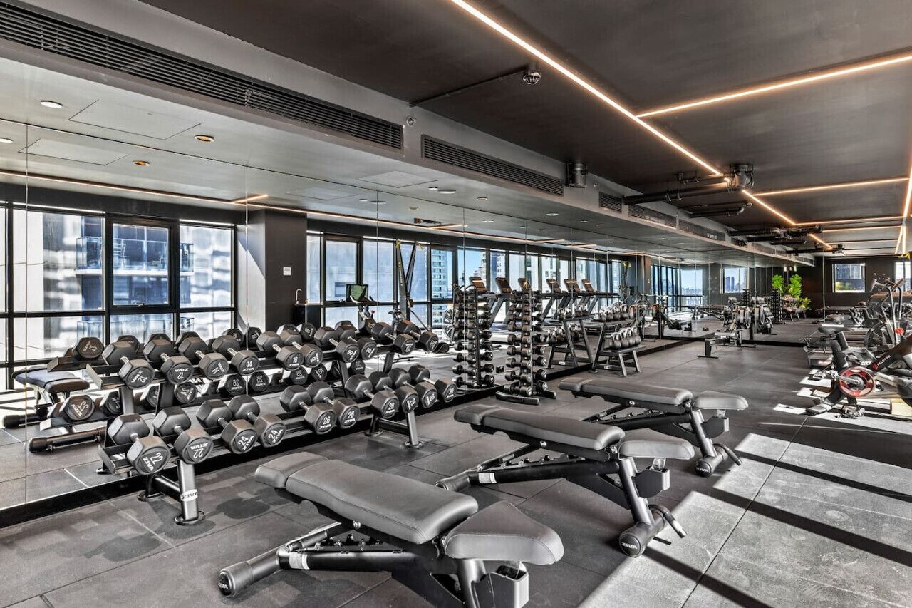 A modern gym with various exercise equipment, large windows, and a high ceiling.