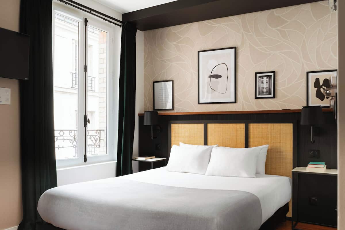 A hotel room with a large bed, a window with curtains, a wall with framed artwork, and a nightstand with a lamp.