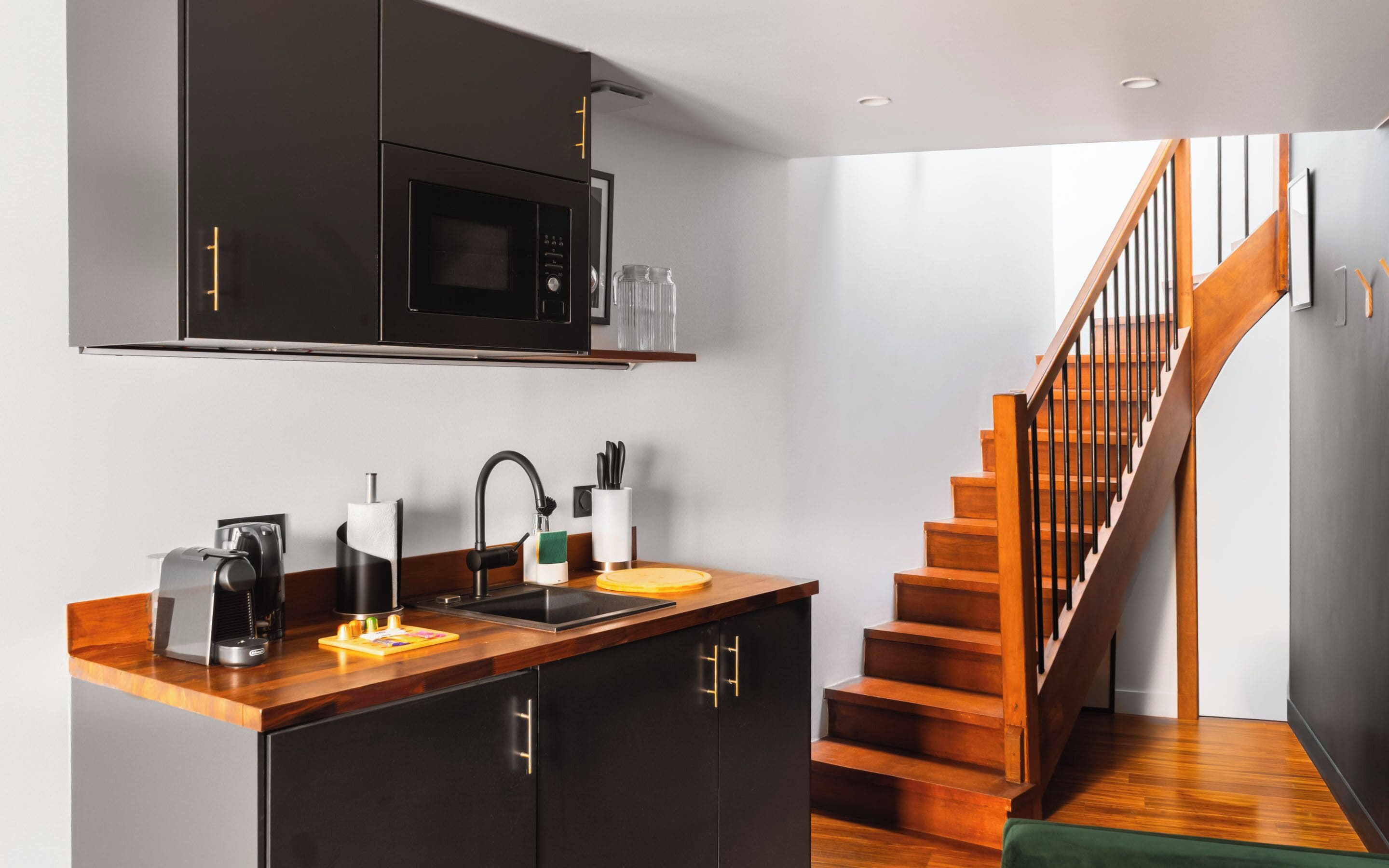 A modern kitchen with a microwave, sink, and wooden countertop. A staircase with wooden steps and a metal railing leads to another floor.