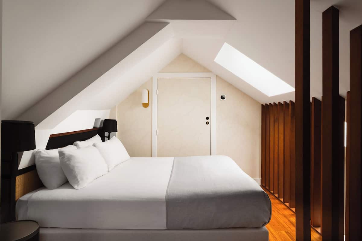A modern bedroom with a skylight, a bed with white and gray bedding, a bedside table with a lamp, and a wooden wardrobe.