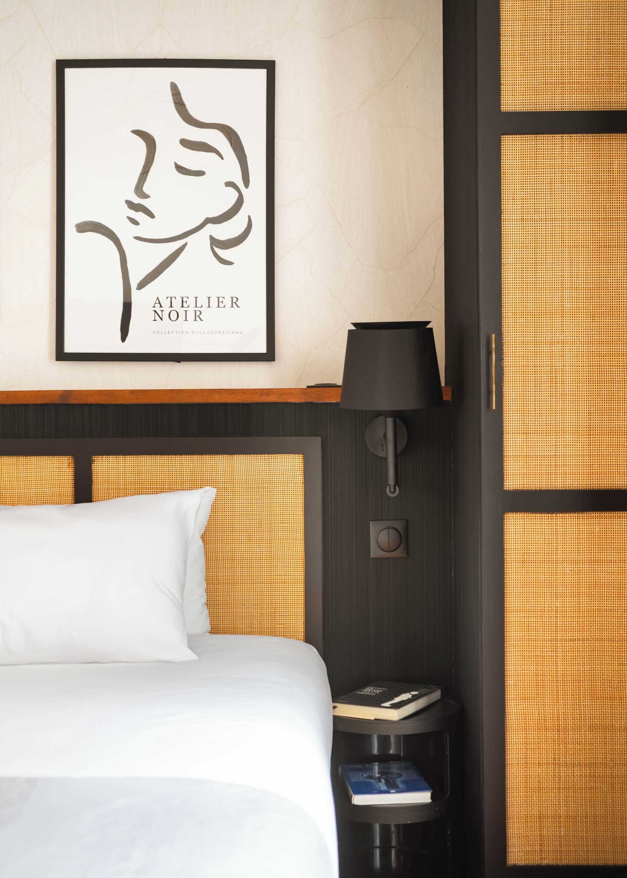 A hotel room with a bed, a framed artwork on the wall, a bedside lamp, and a small table with books.