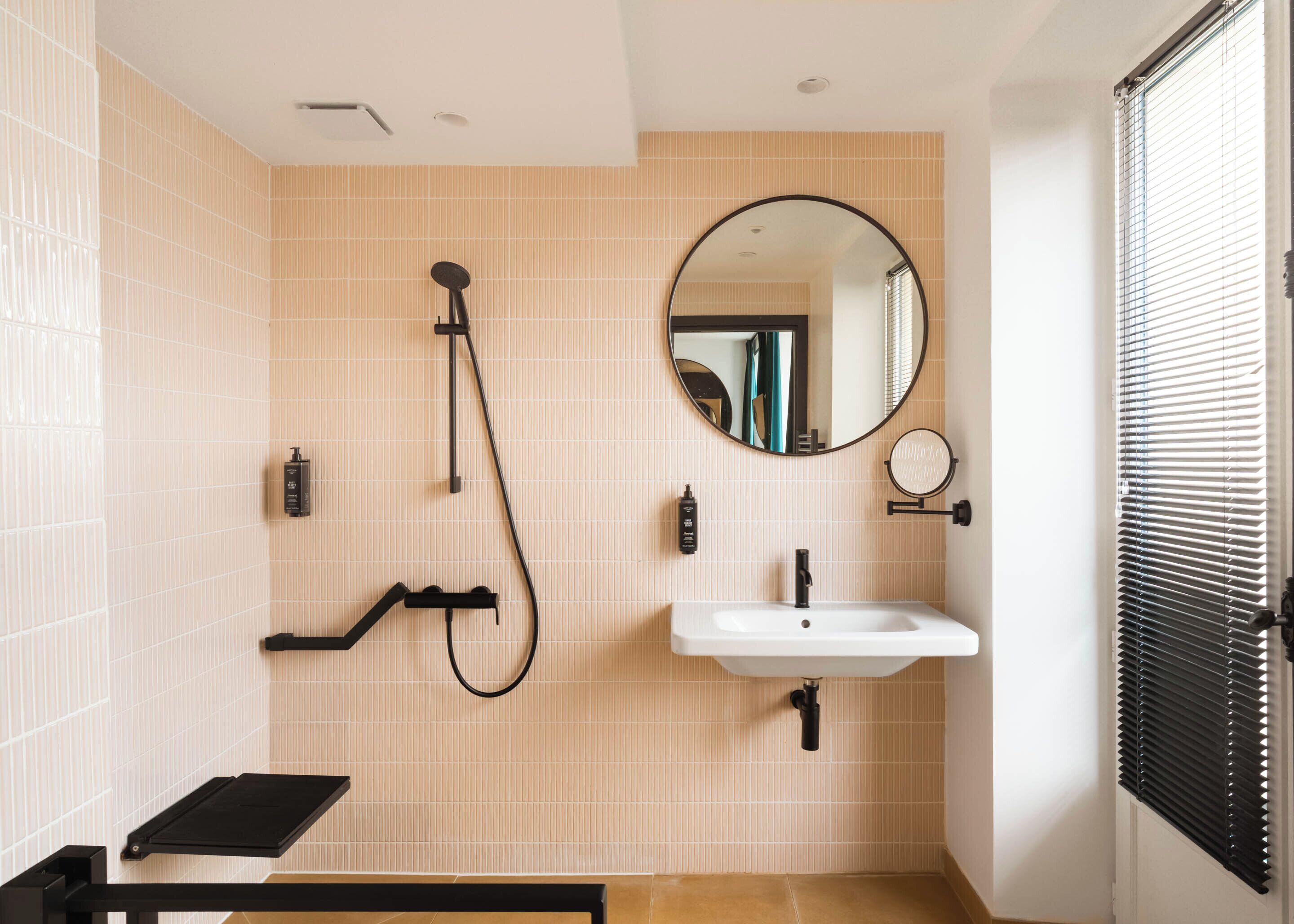 A bathroom with beige tiled walls, a white sink, a black handheld showerhead, a round mirror, and a black towel rack.