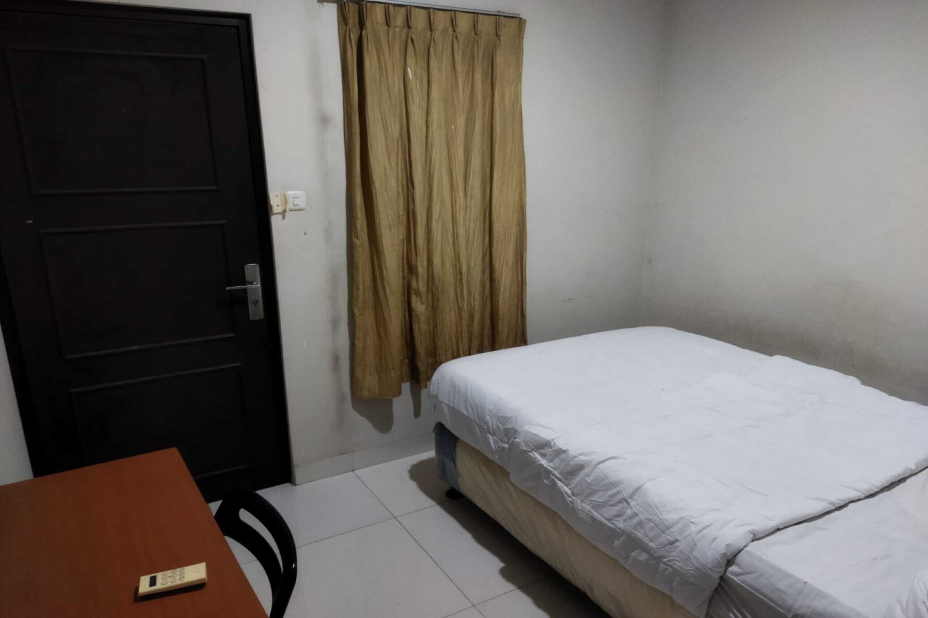 Una habitación pequeña y sencilla con una cama, una mesa con un control remoto, una puerta y una cortina.