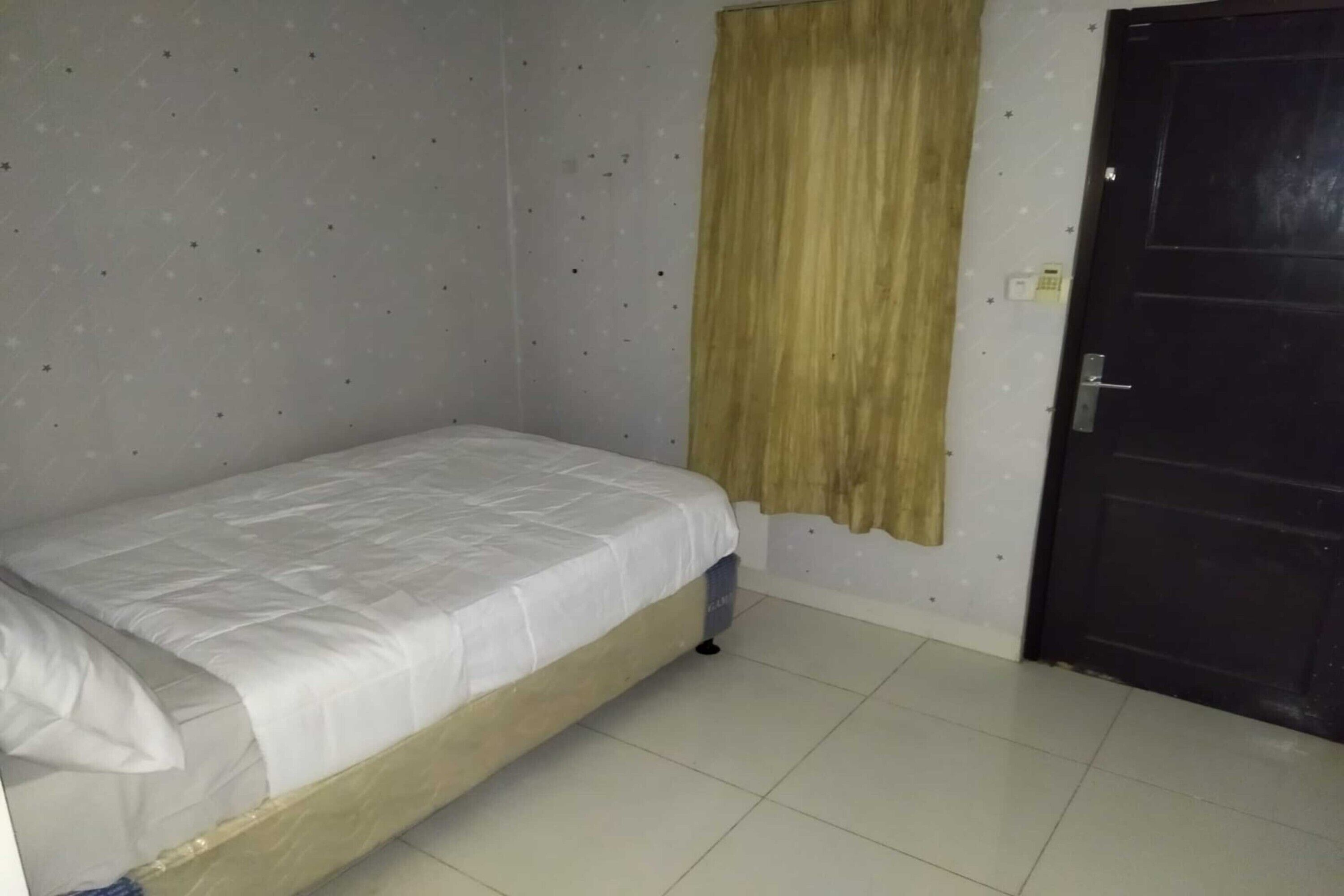 Una habitación pequeña con una cama individual, estructura de madera, colchón blanco sencillo y una cortina en la ventana.