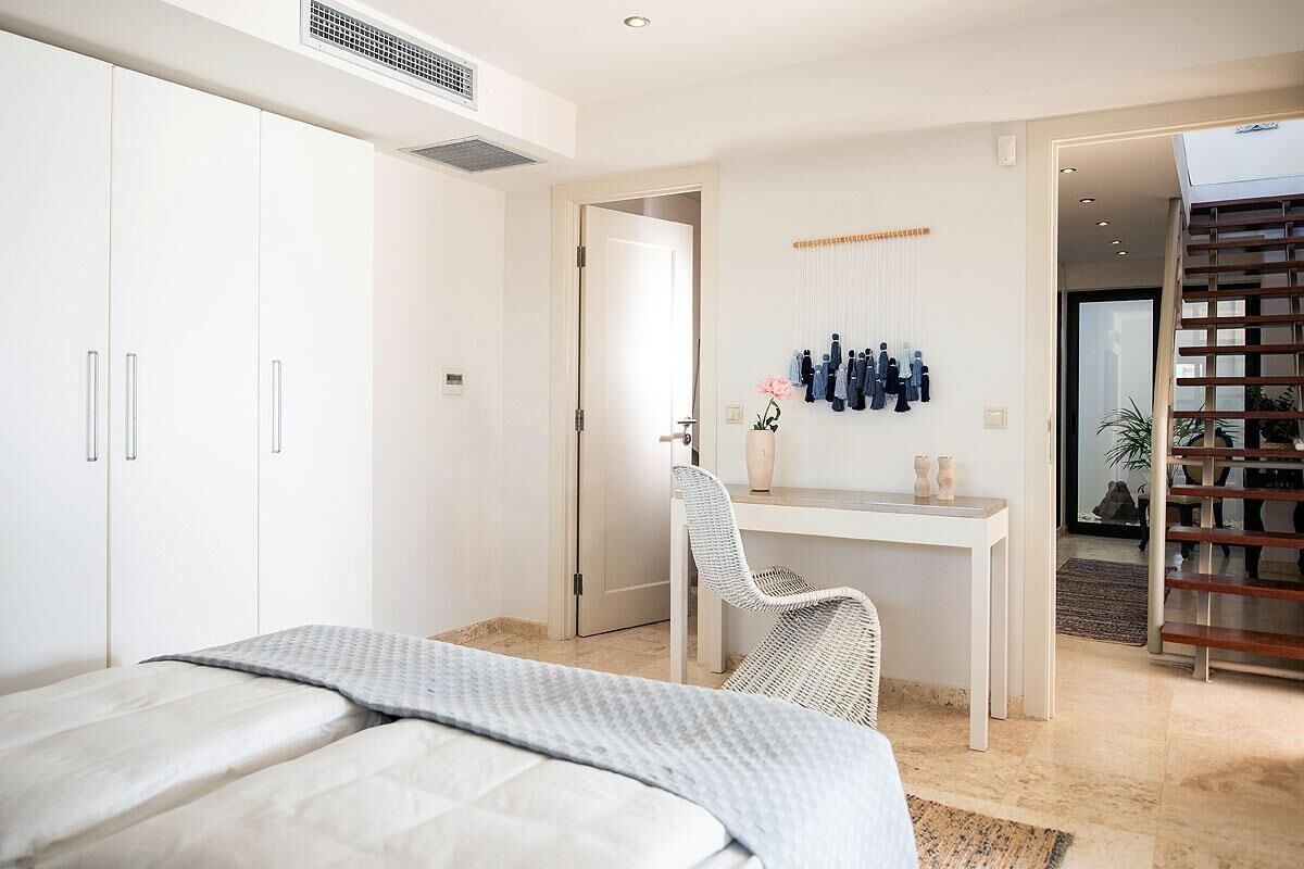 Een slaapkamer met een bed, een bureau en een stoel. Er is een kledingkast, een deur en een plank met flessen.