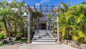 Un chemin menant à un bâtiment doté d’une entrée en forme de pergola, le tout entouré d’une végétation tropicale.
