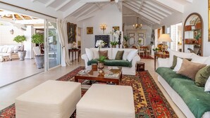 Un salon spacieux avec un plafond haut, un ventilateur de plafond et une grande fenêtre. On y trouve un canapé vert, un canapé blanc et une table basse avec des livres.