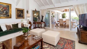 Un salon spacieux avec un plafond haut, un canapé, une table basse et un téléviseur. On aperçoit un coin repas avec des chaises et une table à travers une porte ouverte.