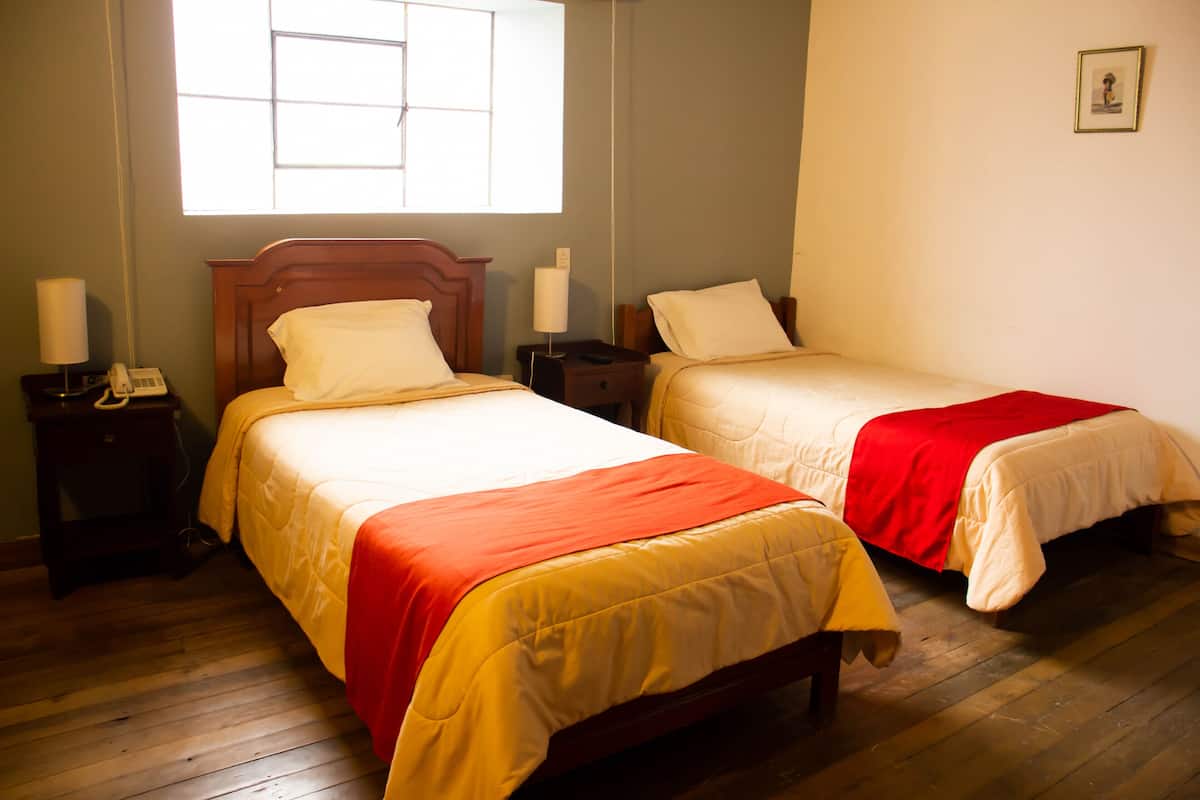 A hotel room with two beds, wooden flooring, a window with blinds, bedside tables with lamps, and a framed picture on the wall.