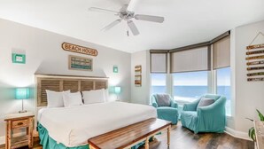 A bedroom with a large bed, a ceiling fan, a view of the ocean, and a sign indicating 'BEACH HOUSE'.