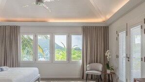 Une chambre à coucher avec un lit, une chaise, une petite table et une plante en pot. Il y a des fenêtres avec des rideaux et une vue sur la verdure à l’extérieur.