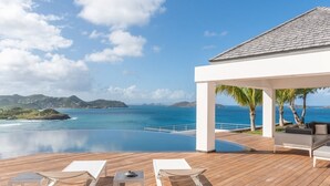 Une terrasse en bois avec des chaises longues blanches et une petite table, d’où l’on peut admirer une mer d’un bleu limpide et des îles lointaines.