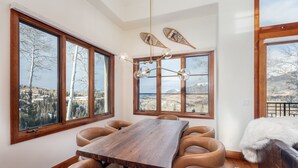 A dining area with a wooden table and leather chairs, large windows with a view of trees and mountains, and a unique leaf-shaped chandelier. - 15 Aspen Ridge-Curate Telluride-Ski In/Out, Hot Tub, Firepit, Walk to Gondola (Mountain Village)