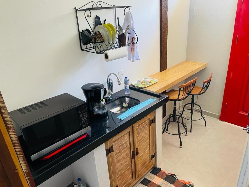 A compact kitchen with a black microwave, a sink, and a wooden countertop. There is a metal rack above the sink holding dishes and a roll of paper towels. A wooden bench and two chairs are positioned near the bench.
