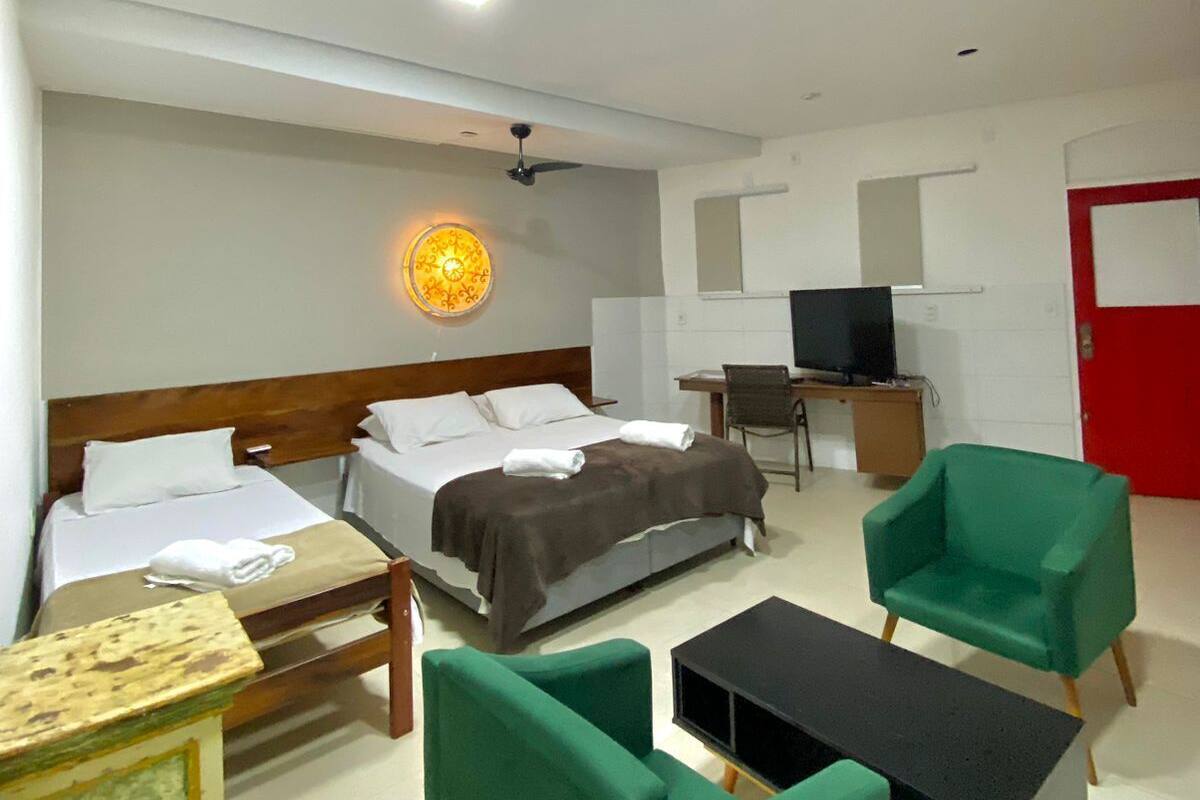 Une chambre d’hôtel avec un lit, deux fauteuils verts, une petite table, un téléviseur et une porte rouge.