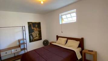 A bedroom with a bed, a small table, a shelf, a painting, and a window.