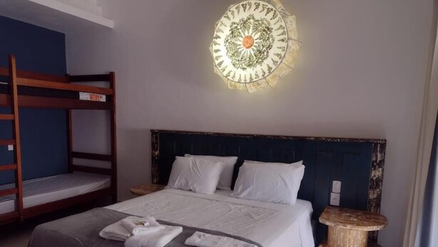 Un dormitorio con una cama litera, una cama grande con ropa blanca, una lámpara de pared redonda y una pequeña mesa redonda con una mesita de noche tejida.